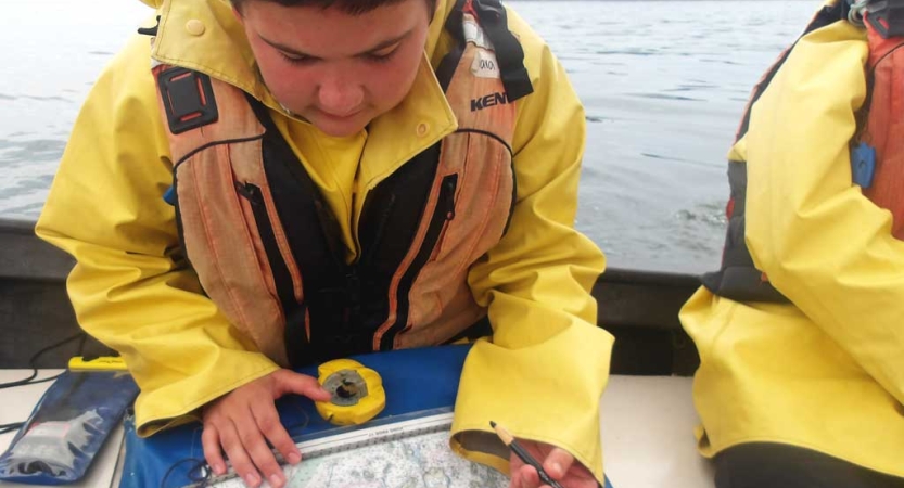 A student wearing a life jacket and yellow raincoat examine a map while sitting on a sailboat. 